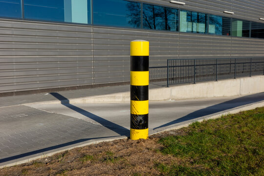 A vertical yellow and black striped safety bollard stands prominently in front of a modern industrial building, marking the edge of a concrete ramp under bright daylight.