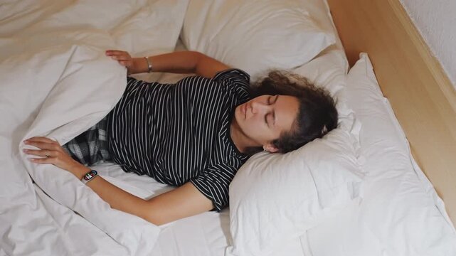 Young woman waking in hotel bed, soft morning light on white duvet, polkadot dress, slow stretch and sigh, bedside wood headboard, tidy pillows, smartwatch on wrist, calm relaxed atmosphere for travel