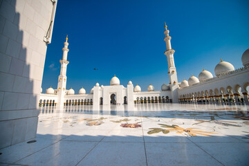 Fototapeta premium Sheikh Zayed Grand Mosque gleams under brilliant sunlight with white marble domes and minarets reflecting intense desert rays against clear blue skies