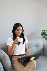 Obraz premium Young woman sitting comfortably on the sofa at home, doing homework or working on laptop while enjoying a cup of coffee, looking relaxed and focused.