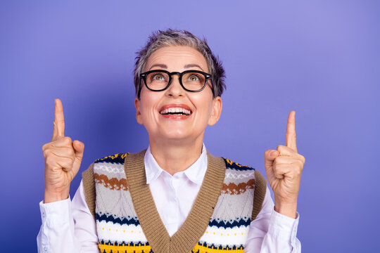Joyful mature woman with glasses and colorful sweater vest smiles and points upward in a studio portrait against a purple background