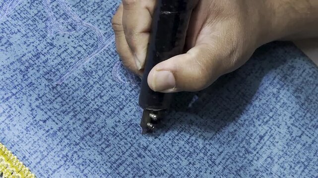 Skilled artisan uses a pyrography tool to engrave Arabic calligraphy on a blue prayer mat, highlighting precise craftsmanship and traditional Islamic textile art in a detailed close-up.