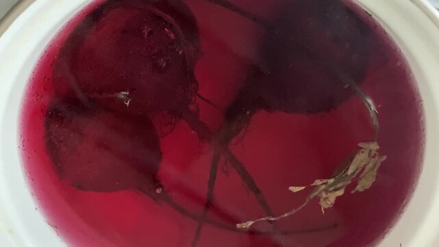 Top-down view of whole fresh red beets boiling in dark red water inside a white cooking pot.