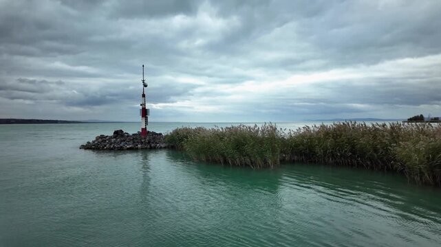 Breakwater with a red tower and reeds and rocks in Tihany, Hungary. Disquiet water surface, waves crashing on the Lake Balaton shoreline on a grey and windy autumn afternoon.