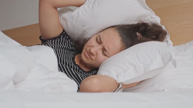 Hotel guest covering ears with pillow, struggle to sleep on white sheets in compact hotel room wristwatch and wooden headboard visible muffled urban noise sparks frustration and restless tossing.