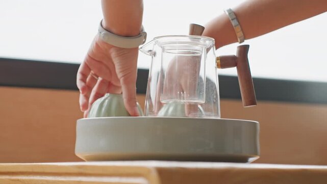 Hotel staff arranging tea tray swiftly on wooden table by window, hands set glass kettle and ceramic cup with careful placement, bracelets and smartwatch visible, focused motion and efficient room