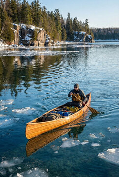 Man paddling a wooden canoe on a frozen lake surrounded by icy water, snow-covered rocks and pine forest in winter wilderness landscape.