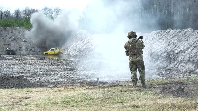 Soldier aiming and firing a handheld grenade launcher at a vehicle in a quarry. Burst of fire and smoke during the shot. Concept of military training, combat readiness, and coordinated unit operations