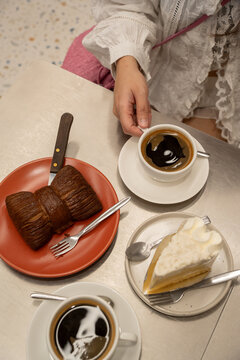 Aesthetic overhead view of a woman's hand holding a black coffee cup next to a chocolate bow-tie croissant and a slice of coconut cream cake on a modern metallic caf&eacute; table.