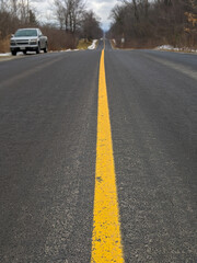 Obraz premium A pickup truck parked to the side of an empty rural road stretching to the horizon, dominated by a bright yellow center line. Bordered by bare winter trees beneath an overcast sky, evoking travel.