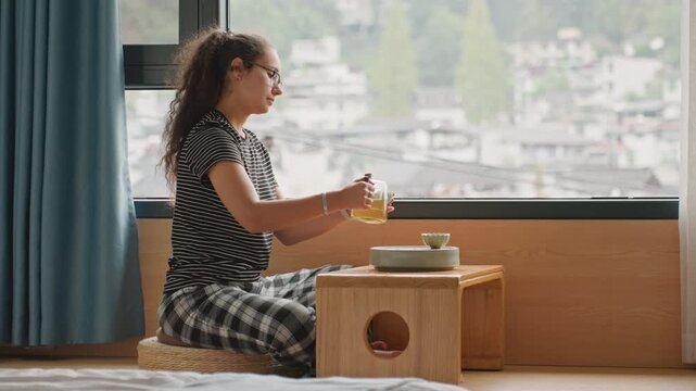 Hotel window tea ritual woman seated by window sipping tea at low table, city view through glass, patterned top and plaid pants, soft curtain, calm reflective mood, solo traveler enjoying slow stay
