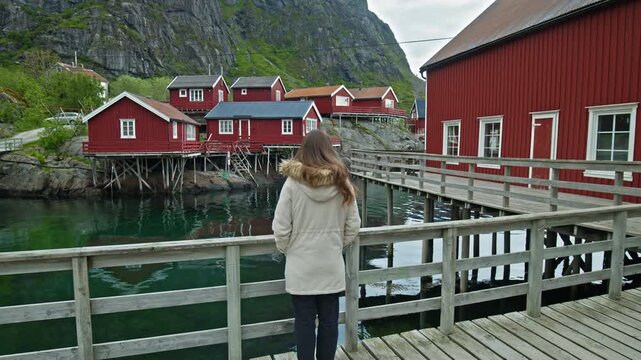 Tourist on pier viewing traditional fishing village in Lofoten, Norway. Red rorbuer cabins and calm fjord with mountain scenery.