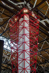 Fototapeta premium Striking red geometric lattice framework encasing tall white cylindrical column, extending upwards into modern industrial-style ceiling with exposed metal beams, trusses, and skylights.
