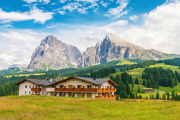 Alpine hotel building on green meadow at Seiser Alm in Dolomites, Italy, with mountain peaks and forest landscape. Ttravel, tourism and hospitality concepts in Alpe di Siusi, Dolomiti, Alto Adige
