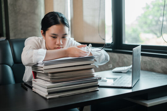 A young woman in a modern office, looking stressed with a large stack of documents and a laptop on her desk.