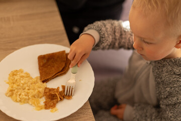 Left handed little child eating pancakes with a fork sitting at the dinner table. Selective focus