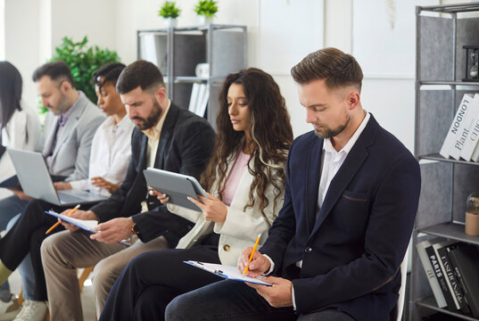 Employees, corporate staff members, applicants sitting in row in office working on diverse devices, writing notes, ideas in clipboard, preparing for job interview or upcoming meeting with clients