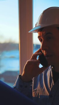 A man in a construction hat speaks on a mobile phone indoors. The man appears focused as he engages in a conversation, with natural light illuminating the scene.