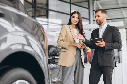 Car salesman using digital tablet discussing specifications with female client at modern auto dealership showroom