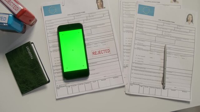 Top view flatlay close-up of EU visa paperwork with red rejected stamp, smartphone with green chromakey screen, passport, on immigration official desk at consulate, no people