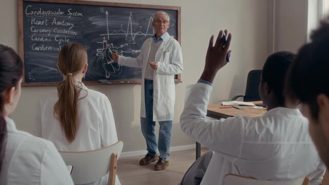 Rear view shot of African American male medical student raising his hand to answer question sitting around classmates in front of Caucasian senior male professor speaking at chalkboard