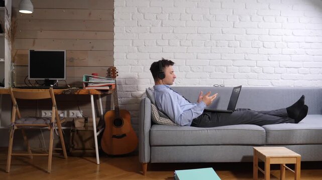 Businessman relaxing on a couch during a video call, wearing headphones and actively participating in a remote work meeting from his comfortable home office setup