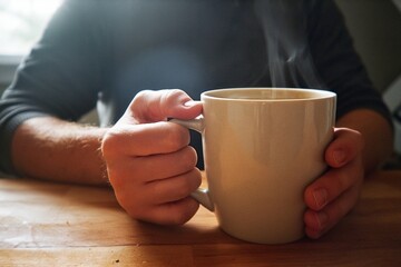 Fototapeta premium Hands holding steaming coffee mug on wooden kitchen counter in morning light