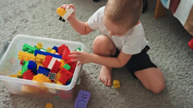 Young son exploring colorful bricks on carpet, reaching into plastic bin to pick and examine pieces, testing fit and texture while sitting barefoot, playful energy and focused curiosity in bright home
