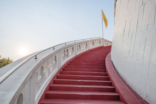 A low-angle view of a red curving staircase with white balustrades leading up the Golden Mount at Wat Saket in Thailand, decorated with waving Thai and Buddhist flags against a clear sky.