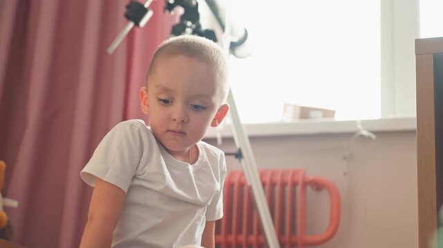 Curious boy climbing into plastic bin, son balancing on edge and peering toward window with soft backlight, colorful bricks in foreground, playful expression and subtle mischief capture independent