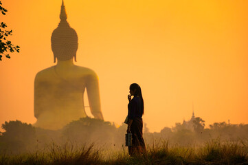 Fototapeta premium Silhouette of Woman Standing in Field with Giant Buddha Statue at Sunrise