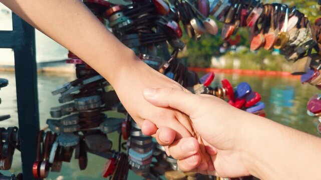 Two people are holding hands by interlacing fingers, expressing love and connection, with many colorful love locks fastened to a bridge railing in the background, signifying eternal partnership