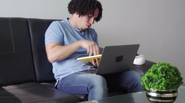 Hispanic man with curly hair studying online, taking notes on a notepad while sitting on the couch