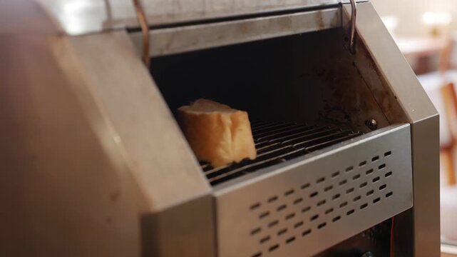 A guest places a slice of bread with metal tongs onto a moving conveyor toaster belt designed for automatic browning in a buffet preparation station. Self service.