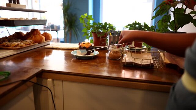 Professional barista carefully taking a delicious chocolate chip muffin from the display case with tongs and placing it on a small plate for a customer in a cozy and modern cafeteria