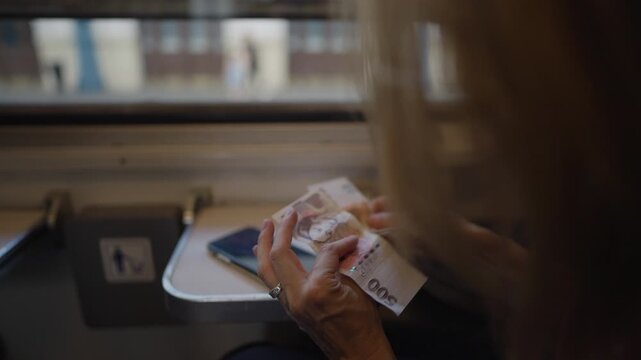Close-up of an unrecognizable elderly woman's hands counting Hungarian forints banknotes while traveling on a train next to a window