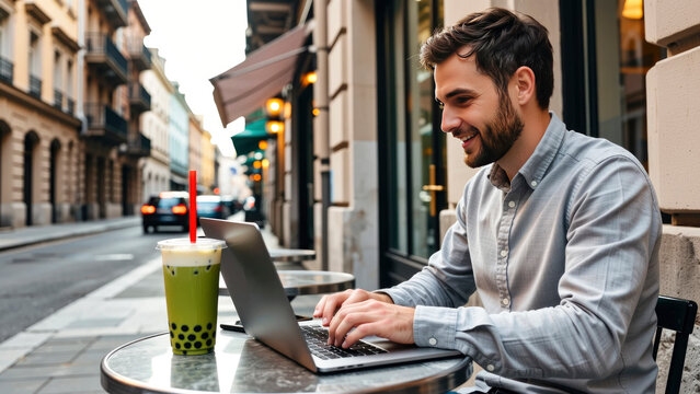 A man sits at a small table outside a cafe, typing on a laptop. Cars pass on the street while he enjoys a drink. The sun shines on the lively urban scene