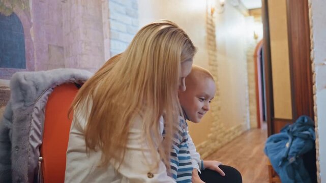 Mom tucks son into warm coat. Playful reassuring scene as mother leans close to fasten zipper and whisper encouragement, child in striped sweater looks up with trust soft hallway light, gentle kiss