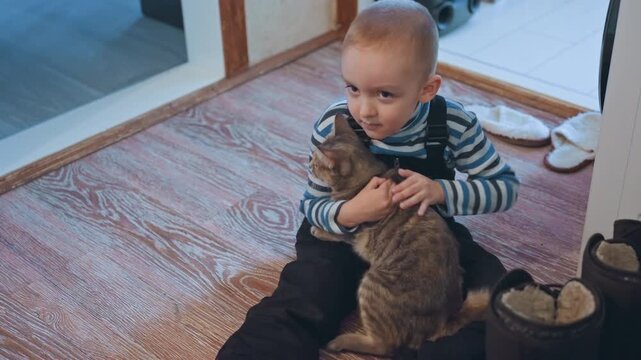 Son hugging tabby cat on rug, striped shirt, seated on floor, warm carpet and slippers nearby, close embrace with purring cat, tender caregiving gesture, quiet domestic afternoon playtime