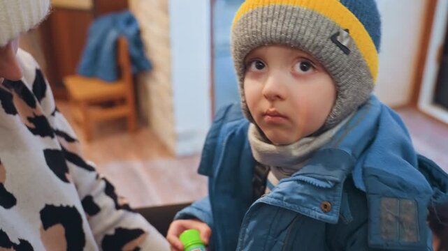 Mom zipping son into coat, close entryway scene with beanie and pompom, attentive hands adjusting zipper, child looking up with quiet trust, warm indoor light, caring family routine