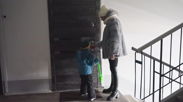 Winter stairwell mom and son enter apartment door, mom hands keys while child holds toy shovel, bundled coats, hat and scarf, quiet stair landing, intimate everyday farewell full of gentle care