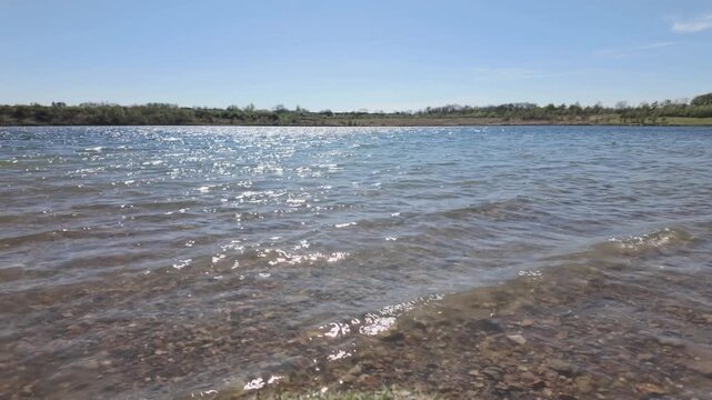 Short sequence at Beeby's Lake West, South Peterborough, Cambridgeshire, England, UK. Waves lapping up at the shore of the lake with the bright blue sky and sun shining reflecting off the water.