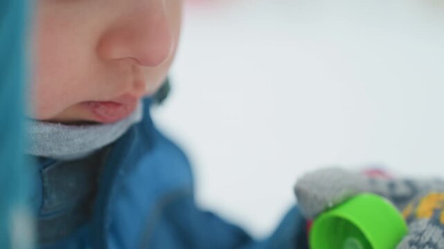 Toddler blowing soap bubble closeup snowy background, blue jacket and knit hat, mittens holding green wand, visible breath and delicate bubble formation, soft shallow depth, candid winter park moment