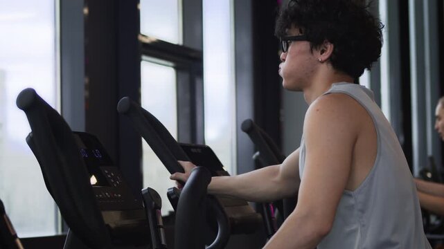 Side view of curly haired young Asian man doing cardio after workout, using elliptical trainer in gym