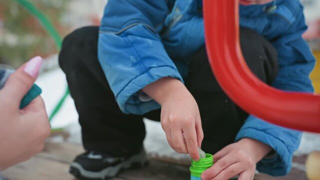 Winter playground mom teaches son bubbles, green bottle and wand, blue jacket and knit hat, careful hands and crouched posture, soap suds catching light, playful joyful outdoor moment
