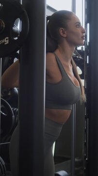 Vertical side view shot of young Caucasian woman focused on squatting in smith machine during training in gym