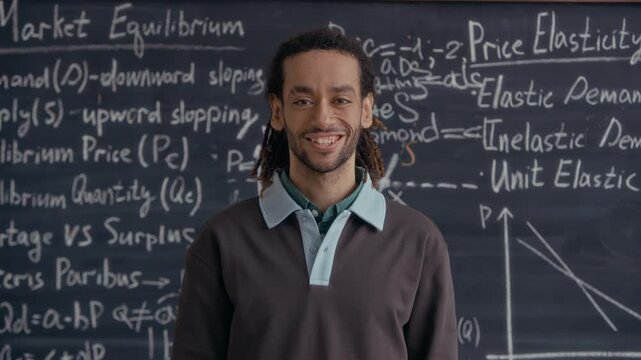 Medium eye level portrait of Biracial male student with dreadlocks standing in front of chalkboard with economic graphs and formulas in classroom