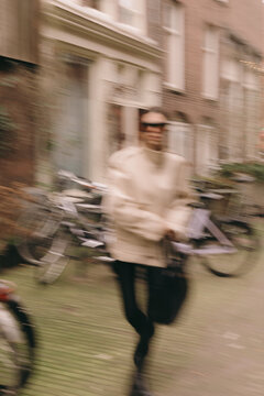 Artistic extreme motion blur of stylish woman in beige jacket and sunglasses walking fast on city street with bicycles, dynamic urban rush, cinematic movement effect, modern aesthetic, blurred
