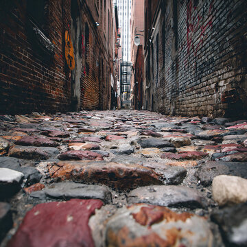 Photograph of a narrow, cobblestone alleyway between tall, weathered brick buildings, with graffiti on the walls and a clear view of tall skyscrapers in the distance.