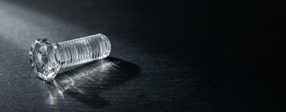 Transparent glass bolt lying on a dark surface with dramatic shadow, industrial object concept, minimal design, precision and engineering, modern material.